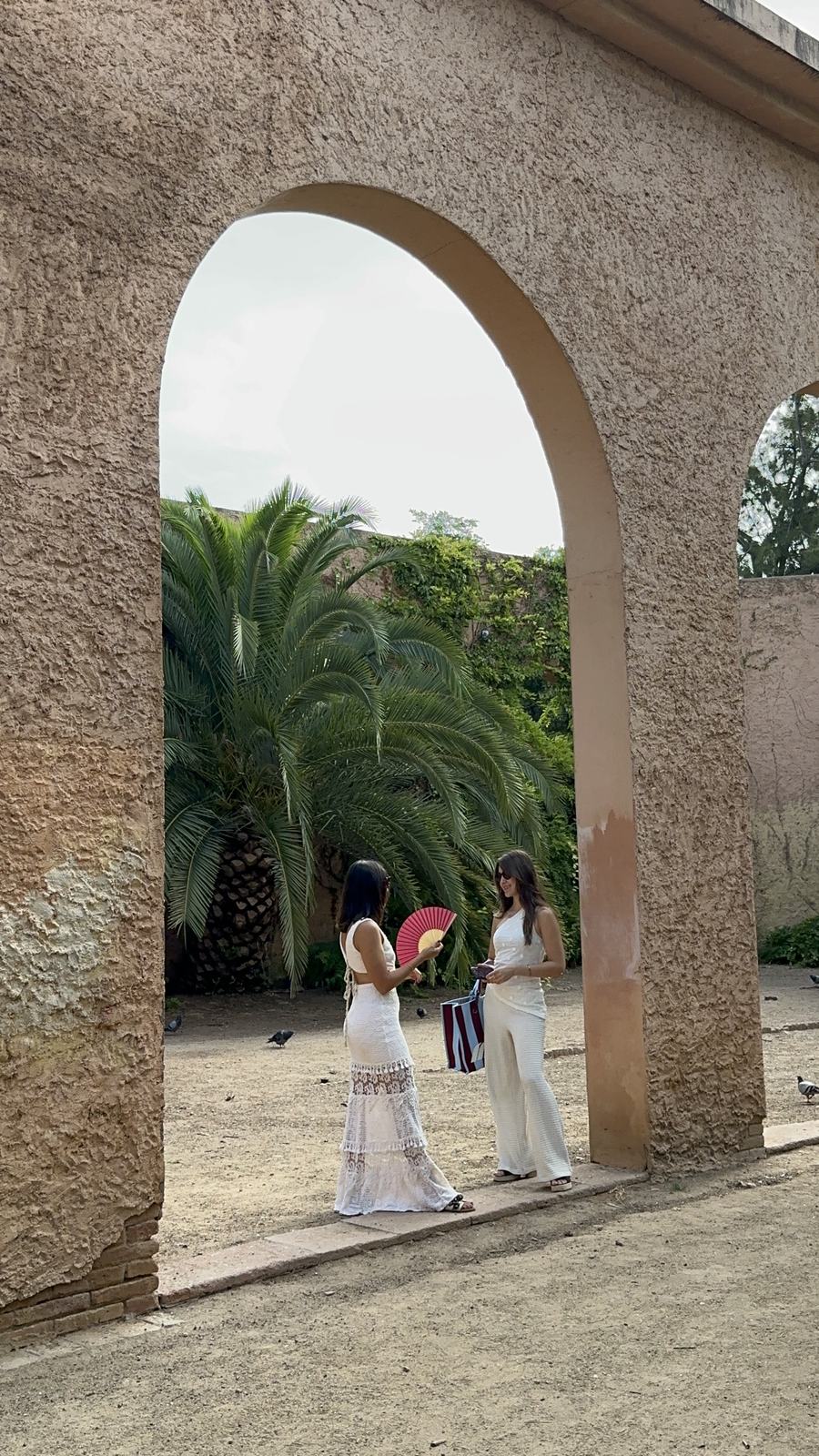 Two women standing together under a stone archway beside a tall palm tree.