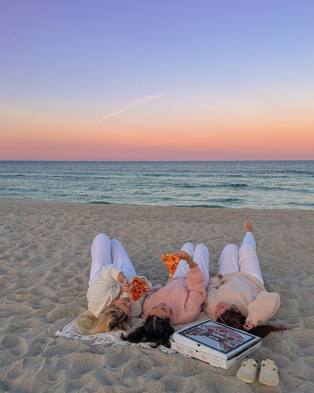 Three women lying together on a beach at sunset, sharing a pizza.