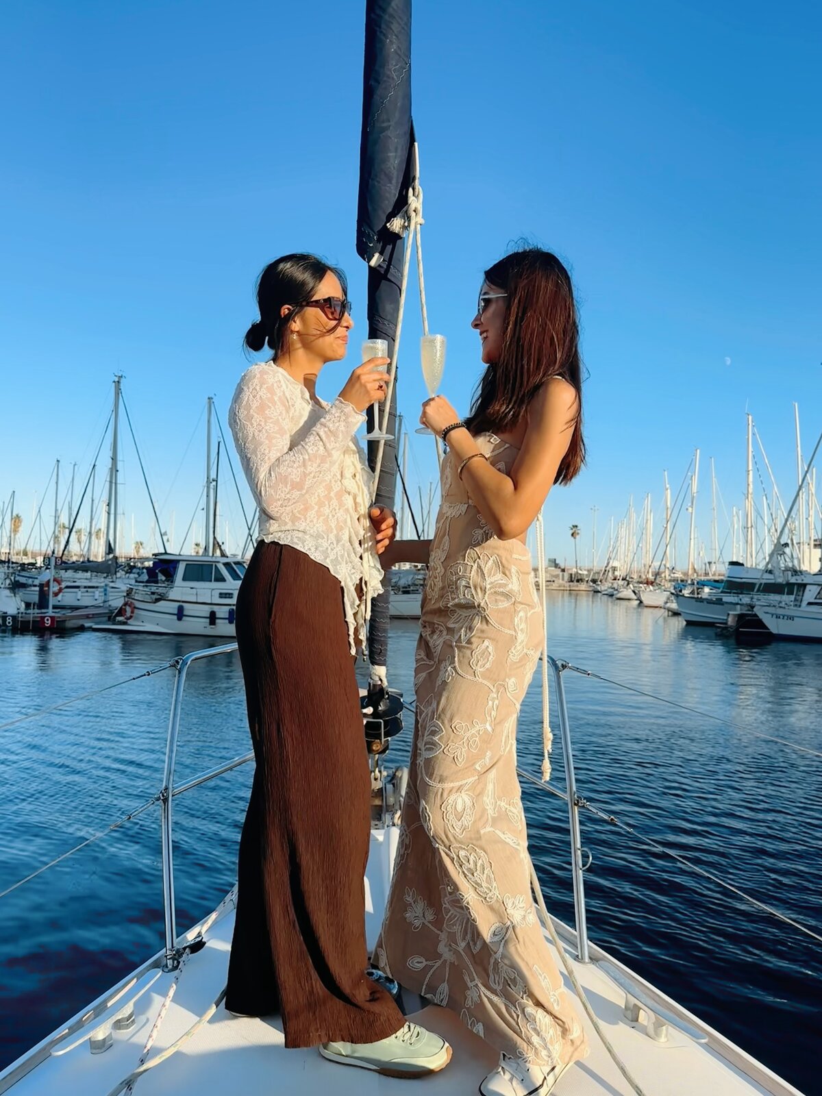 Two women in elegant dresses toasting champagne on a sailboat bow in a marina at golden hour.