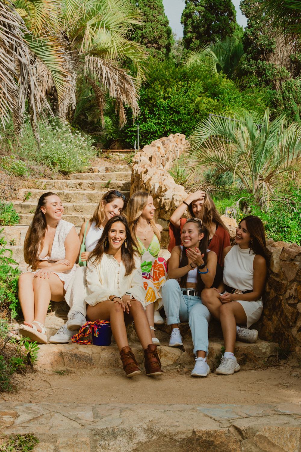 A group of women laughing together on stone garden steps lined with palm trees.