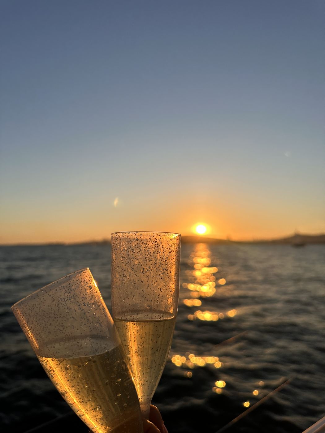 Two glittering champagne flutes raised toward the setting sun over open water.