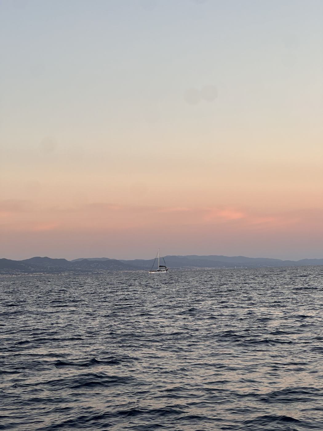 A lone sailboat silhouetted against coastal hills under a pastel sunset.
