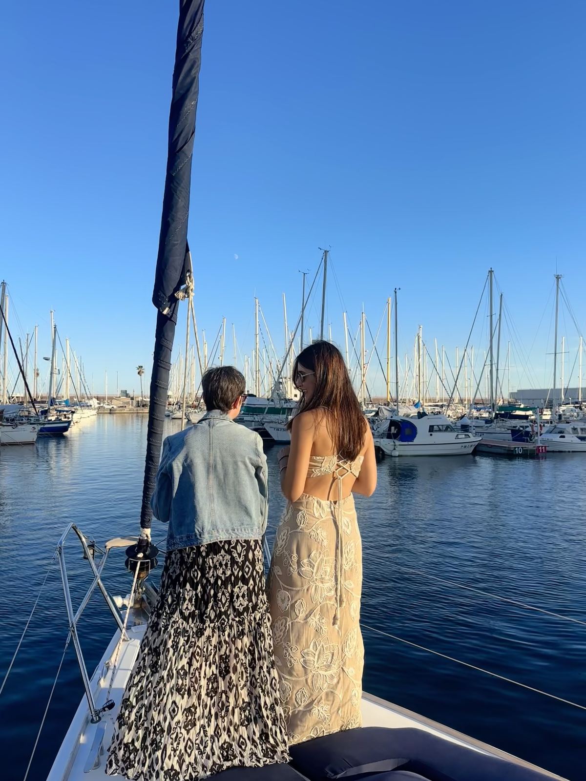 Two women, backs to the camera, in elegant dresses, walking along a marina full of sailboat masts at golden hour.