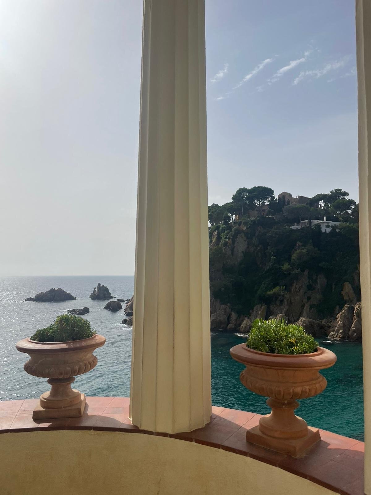 A stone balcony with potted plants overlooking a pine-covered Costa Brava cove.