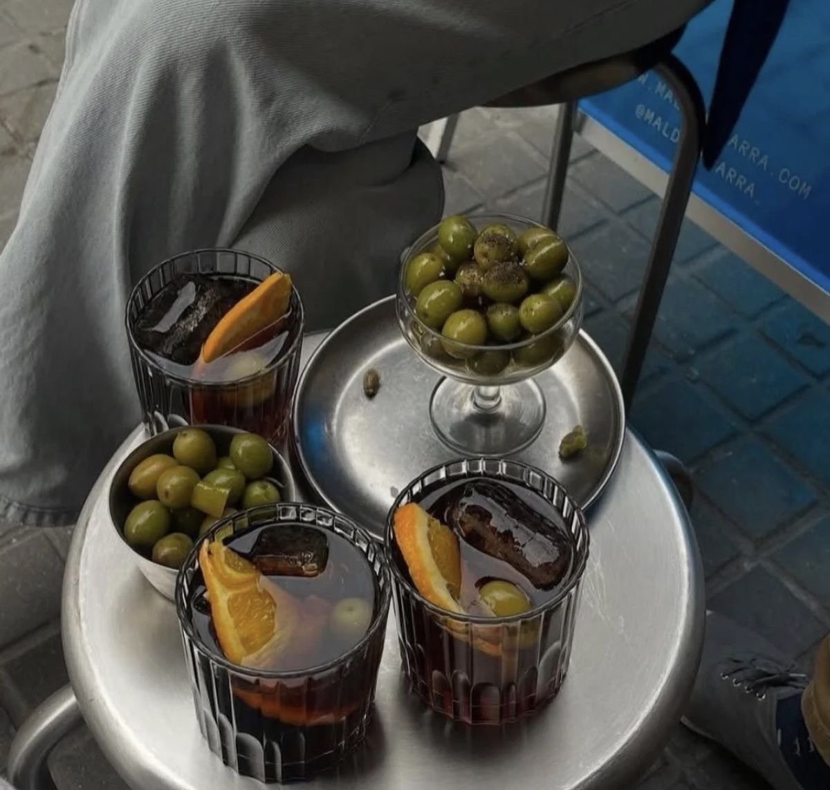 Glasses of vermouth with orange slices and ice, served alongside bowls of green olives on a street table.