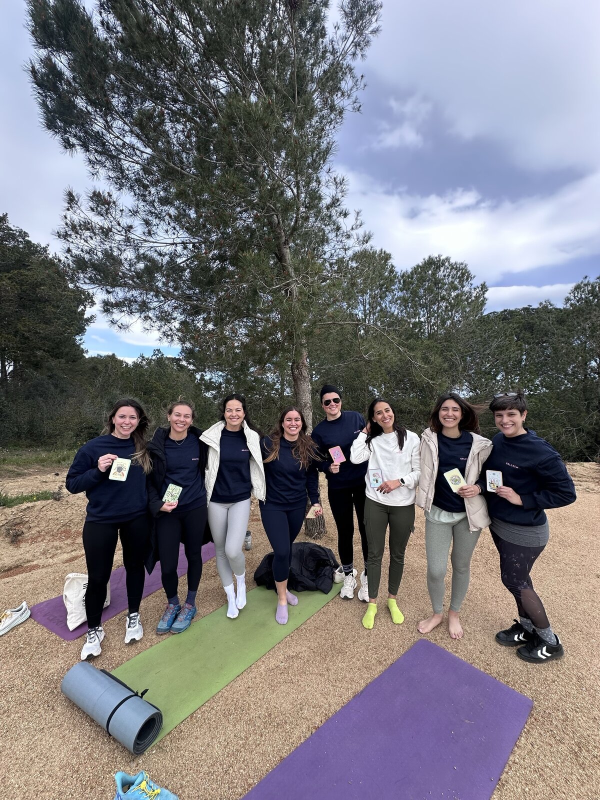 A group of women on yoga mats together outdoors under a pine tree.